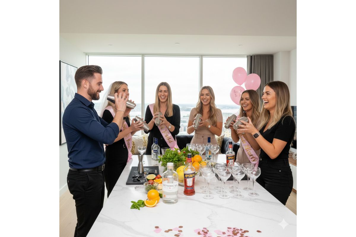 A professional male mixologist leading a mobile cocktail making class in Cardiff for a hen party. A group of women in a modern apartment are laughing while learning to use cocktail shakers at a kitchen island stocked with premium spirits and fresh fruit